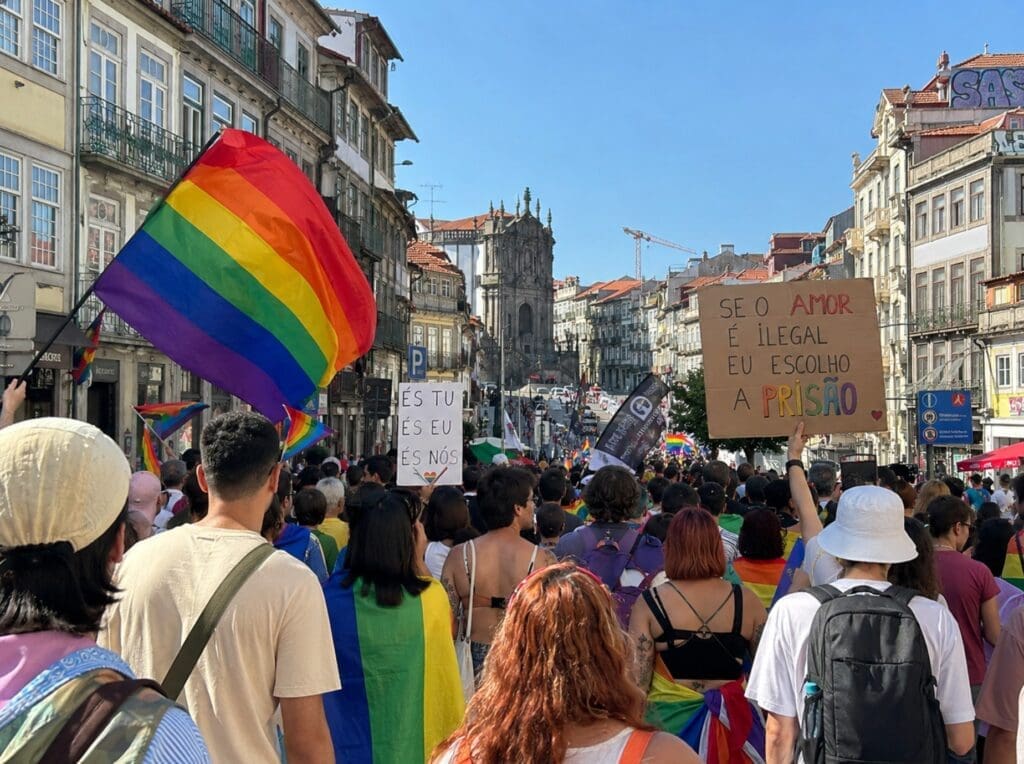 Marcha do Orgulho no Porto vista de trás, com bandeiras, cartazes e arquitetura da cidade ao fundo
