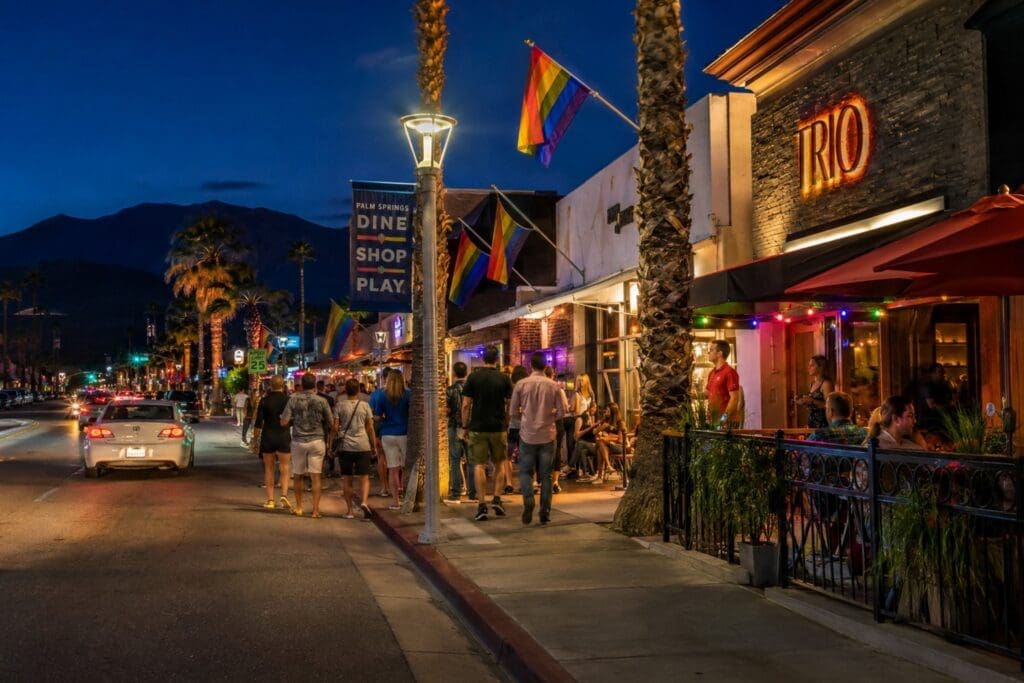 evening photograph of Arenas Road showing illuminated patios, rainbow flags, palm trees, and groups of friends walking between venues