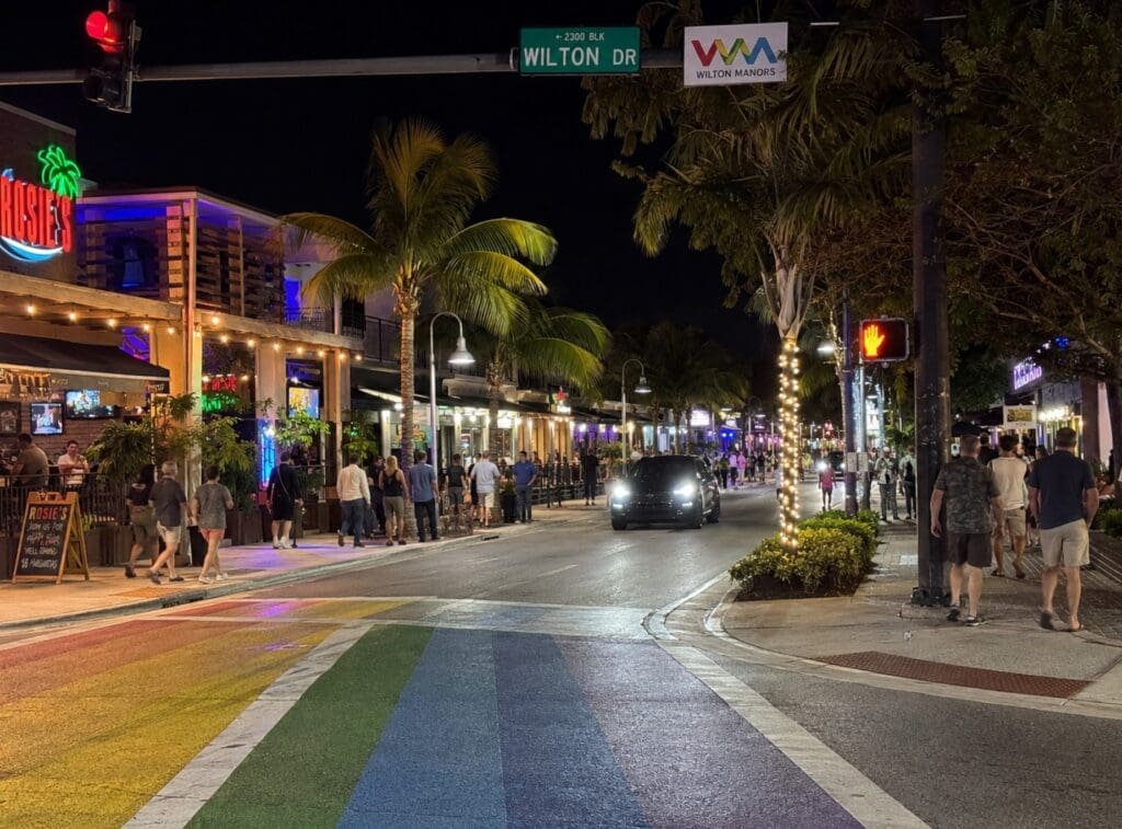 nighttime photograph of pedestrians, illuminated storefronts, and rainbow crosswalks along Wilton Drive