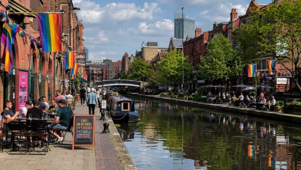 photograph of Canal Street during daytime, with rainbow flags, the canal, and outdoor seating (manchester)