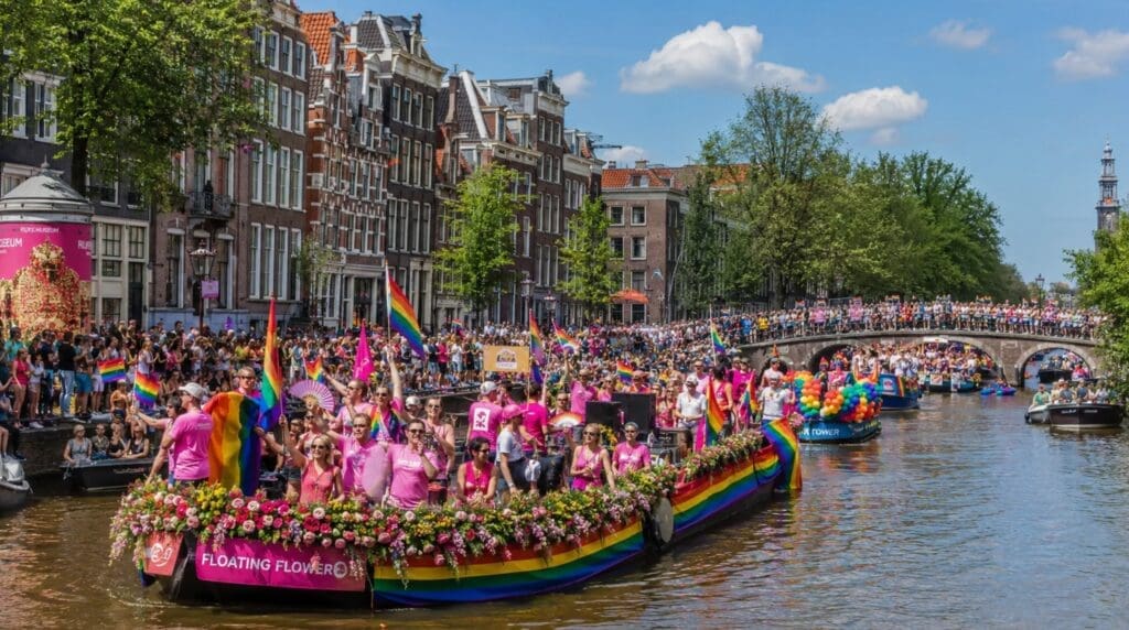 photograph showing an Amsterdam canal during Pride, with decorated boats, historic houses, and spectators along the waterfront