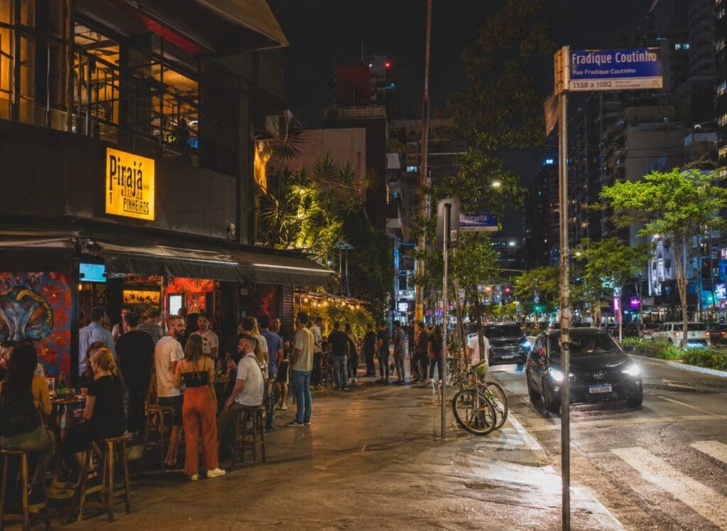 uma fotografia de uma rua movimentada de Pinheiros à noite, com bares, luzes urbanas e pessoas vistas de longe